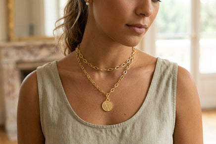 A woman wearing a layered gold necklace set, featuring a delicate chain and a longer chain with a circular coin pendant, paired with a light beige linen tank top.